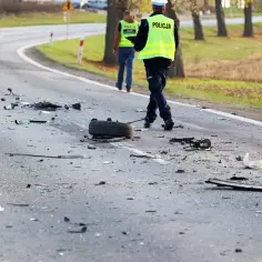 Na drodze po kolizji leżą szczątki, opona i elementy pojazdów. Policjanci w kamizelkach odblaskowych zabezpieczają miejsce zdarzenia, badając okoliczności ucieczki z miejsca kolizji.