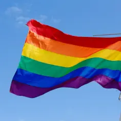 Tęczowa flaga powiewa na maszcie na tle błękitnego nieba. Kolory symbolizują różnorodność i dumę społeczności LGBT.