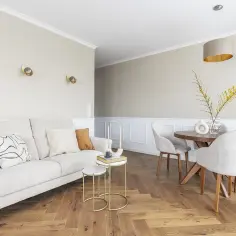 Jasny salon z sofą, stolikami i krzesłami przy okrągłym stole. Wnętrze zdobią białe listwy przypodłogowe, dodając elegancji.