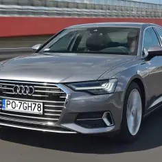 Nowe Audi A6 w kolorze szarym jedzie po drodze. Kierowca w okularach prowadzi luksusowy samochód.