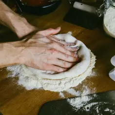 Dłonie formujące ciasto na pizzę na drewnianym blacie obsypanym mąką.