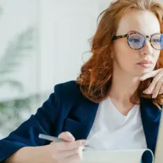 Kobieta w okularach pracuje przy laptopie. Zastanawia się nad dokumentami, być może nad wnioskiem o refundację okularów przez pracodawcę.