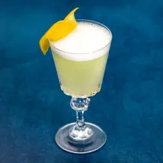 Elegancki drink White Lady z pianką i skórką cytryny na tle granatowego blatu.