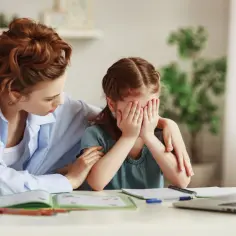 Mama pociesza płaczące dziecko, które nie chce chodzić do szkoły. Dziewczynka zakrywa twarz dłońmi, a mama obejmuje ją ramieniem.