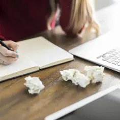 Osoba pisze w notatniku, przygotowując recenzję książki. Obok leży laptop i pogięte kartki.