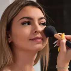 Młoda kobieta z długimi blond włosami nakłada puder pędzlem. Jej makijaż jest subtelny, podkreślający naturalne piękno, w stylu clean girl.