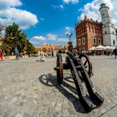 Sandomierz rynek z armatą w tle ratusza i kamienic. Ludzie spacerują po brukowanym placu w słoneczny dzień.