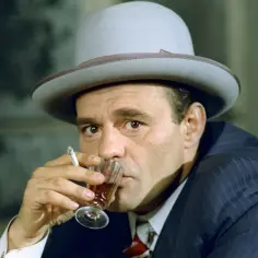 Nikodem Dyzma w kapeluszu, popijający drinka i palący papierosa. W jego oczach widać pewność siebie i przebiegłość.