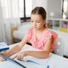 Dziewczynka korzysta z tabletu do nauczania domowego w podstawówce. Na biurku leżą książki i zeszyt.