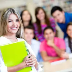 Uśmiechnięta studentka z zielonym zeszytem, gotowa do rozwiązywania zadań z biologii. W tle grupa innych studentów.