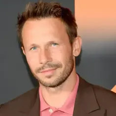 Mateusz Banasiuk, znany z wielu polskich filmów, w eleganckim brązowym garniturze i różowej koszuli.