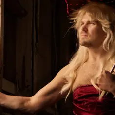 Transwestyta w blond peruce i czerwonej sukience, trzymający parasolkę, patrzy przez okno.