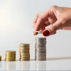 Dłoń z czerwonym lakierem dokłada monetę do rosnącej wieży. Zastanawiasz się, jaka podwyżka przy przedłużaniu umowy czeka Cię w przyszłości?