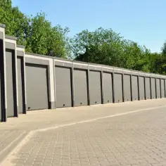Rząd nowoczesnych, szarych garaży betonowych prefabrykowanych, każdy z numerem, stoi wzdłuż utwardzonego podjazdu, otoczony zielenią.