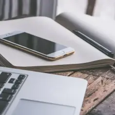 Laptop, telefon i otwarty notatnik z długopisem na drewnianym stole. Idealne miejsce, by zacząć zastanawiać się, jak uzupełnić dziennik praktyk.
