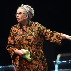Krystyna Janda w barwnej sukni i chustce na głowie, gestykuluje na scenie, trzymając w dłoni chusteczkę.
