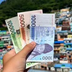 Dłoń trzyma banknoty 1000, 5000 i 10000 wonów. W tle kolorowe domy, symbolizujące ceny w Korei Południowej.