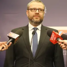 Marcin Wojciechowski w okularach, w garniturze, otoczony przez mikrofony mediów, udziela wywiadu.