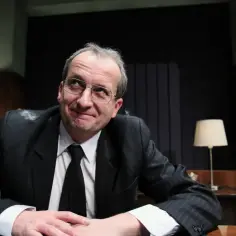 Robert z Kabaretu Moralnego Niepokoju w garniturze, z okularami, zamyślony, siedzi przy biurku.