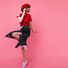 Dziewczyna w stylu pin up w czerwonym beretce i sukience w grochy tańczy na różowym tle.
