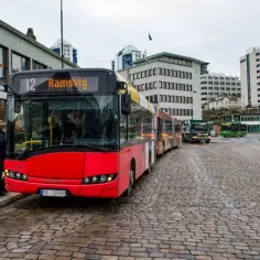 Czerwony przegubowy autobus z numerem 12 "Ramsvig" na wyświetlaczu, stojący na brukowanej ulicy w mieście.