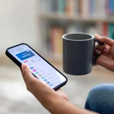 Dłoń trzyma smartfon z aplikacją bankową, gdzie widać dostępne środki i wydatki. Obok kubek kawy. Idealny moment na sprawdzenie swojego subkonta.