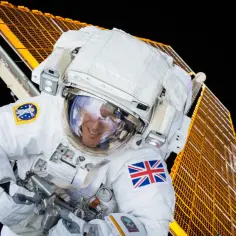 Uśmiechnięty astronauta w białym skafandrze kosmicznym, z brytyjską flagą na ramieniu, pracuje przy panelach słonecznych stacji kosmicznej.