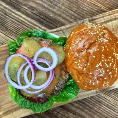 Jak przyprawić mięso wołowe na burgery? Soczysty burger z sałatą, ogórkiem, cebulą i sezamową bułką na drewnianej desce.