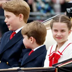 Dzieci Williama i Kate w karocy: książę George, książę Louis i księżniczka Charlotte, uśmiechnięta Charlotte w białej sukience z czerwoną kokardą.