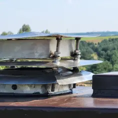 Metalowy daszek na komin, wielopoziomowy, z widocznymi śrubami i nakrętkami. Obok stoi brązowy, kwadratowy element. To inspiracja, jak zrobić daszek na komin.