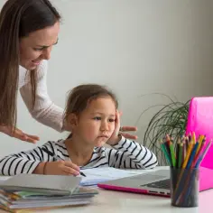 Zestresowana dziewczynka z zeszytem i laptopem, podczas gdy mama z niezadowoloną miną tłumaczy jej coś o ocenach w szkole.