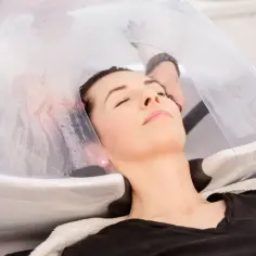 Relaks w trakcie zabiegu hair spa. Kobieta z zamkniętymi oczami leży w fotelu fryzjerskim, pod specjalną kopułą.