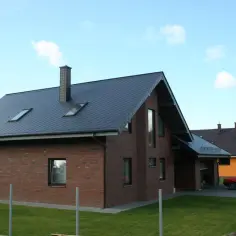 Nowy dom z cegły, z ciemnym dachem i kominem o imponującej wysokości. W tle inne domy i czerwony samochód.