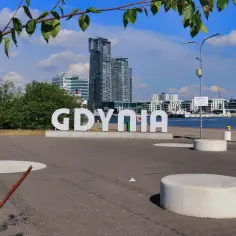 Napis GDYNIA na tle nowoczesnych budynków i żaglowca.