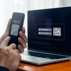 Ręce trzymają telefon z napisem "SCAN TO LOGIN", obok leży laptop z widocznym kodem QR.