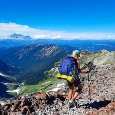 Kobieta z plecakiem wspina się po kamienistym szlaku, podziwiając widok na góry i ośnieżony wulkan. To fragment Pacific Crest Trail.