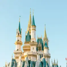 Bajkowy zamek z zielonymi dachami i złotymi detalami, przypominający ten z pierwszego Disneylandu, na tle błękitnego nieba.