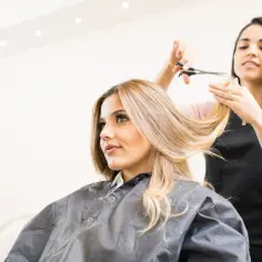Fryzjerka przycina blond włosy klientki, tworząc nową, modną fryzurę. Efekt końcowy to ładnie obcięte włosy.