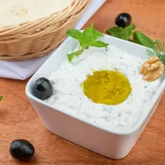 Kremowy sos tzatziki w białej miseczce, ozdobiony oliwą, orzechem i ziołami. Obok oliwki i koszyk z pieczywem.