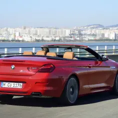Czerwony kabriolet BMW F12 z brązowym wnętrzem jedzie po moście, w tle widać miasto i wodę.