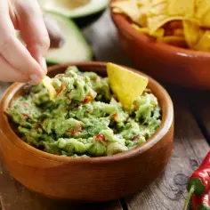 Dłoń zanurza chipsa w misce z guacamole, obok chili i nachosów.