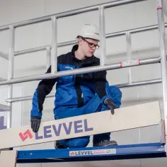 Mężczyzna w kasku i niebieskim kombinezonie demonstruje, jak złożyć rusztowanie aluminiowe, umieszczając drewnianą deskę.