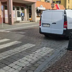 Biały van stoi na brukowanej ulicy, blokując przejście dla pieszych. Obok widać budynek z wejściem.