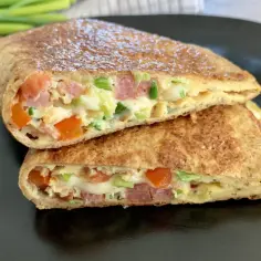 Keto tortilla przepis: złocista tortilla z jajkiem, szynką, pomidorem i szczypiorkiem, pokrojona na pół, na czarnym talerzu.