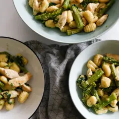 Pyszne gnocchi ze szparagami i kurczakiem w sosie. Danie podane w trzech miseczkach, obok szklanki z wodą i limonką.