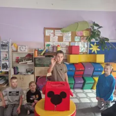 Chłopiec trzyma żołądź, symbol jesieni, przed pudełkiem z Myszką Miki. Dzieci patrzą z zaciekawieniem.