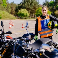 Dziewczyna w kamizelce z literą "L" i kaskiem w ręku gotowa do jazdy na motocyklu. Idealny motocykl na egzamin kat A czeka!