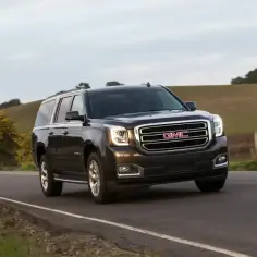Ciemny GMC Yukon XL jedzie po wiejskiej drodze, otoczony zielonymi wzgórzami i drzewami.