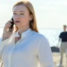 Sarah Snook w białej koszuli rozmawia przez telefon na plaży. W tle widać dwie postacie.