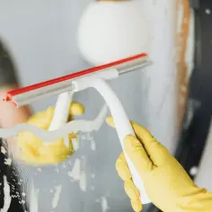 Ręka w żółtej rękawiczce używa ściągaczki do mycia lustra, co może być jednym z najgorszych nawyków podczas sprzątania, przez co dom brudzi się szybciej.